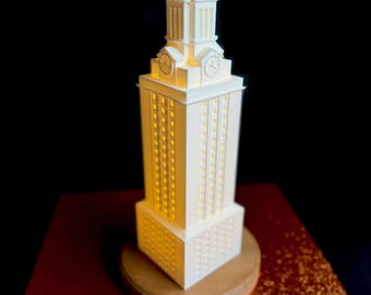 May include: A white 3D printed model of the University of Texas at Austin's iconic tower, the UT Tower, with a clock face and illuminated windows. The model is on a wooden base.