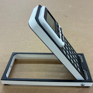 May include: A white and black calculator stand featuring a rectangular base frame and an angled support panel. The calculator is securely held at an incline, displaying a digital screen and a grid of circular buttons on a black faceplate.