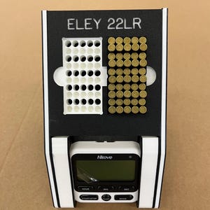 May include: A black and white device with the text "ELEY 22LR" at the top. It features a white grid for holding items, and a digital timer labeled "Nkove" at the bottom. The timer has buttons for hour, min, sec, start/stop, clear, and mode.