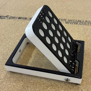 May include: A black and white angled stand with a grid of oval holes. The stand has a rectangular base and a slanted top section. The top section has the text "SER" on it. The stand is on a wooden surface.