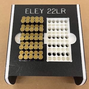 May include: A black and white ammunition holder with the text "ELEY 22LR". The holder contains brass-colored bullets on the left and empty white holes on the right. The holder is designed to hold 50 rounds.