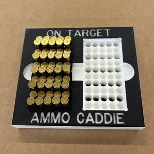 May include: Black and white ammo caddie with the words "ON TARGET" and "AMMO CADDIE" printed on it. The caddie holds several gold-coloured bullets in a rectangular section and has a white section with multiple holes.