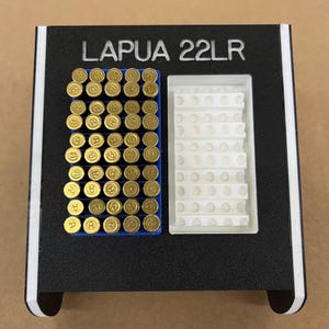 Puede incluir: Un soporte de munición en blanco y negro con el texto "LAPUA 22LR". El soporte tiene un compartimento azul lleno de balas doradas y un compartimento blanco con un patrón de rejilla.