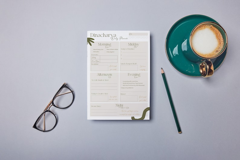 May include: A white daily planner with the title "Dinacharya Daily Planner" and sections for Morning, Midday, Afternoon, Evening, and Night. The planner is on a gray surface with a green pencil, a cup of coffee, and a pair of glasses.