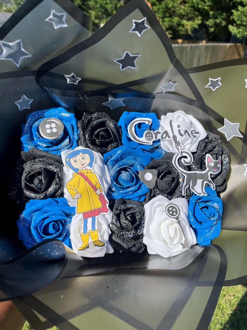 May include: A bouquet of artificial roses in blue, black, and white, with decorative accents. The bouquet includes a character cutout, a button, a key, and the word "Coraline". The bouquet is wrapped in black cellophane with star accents.