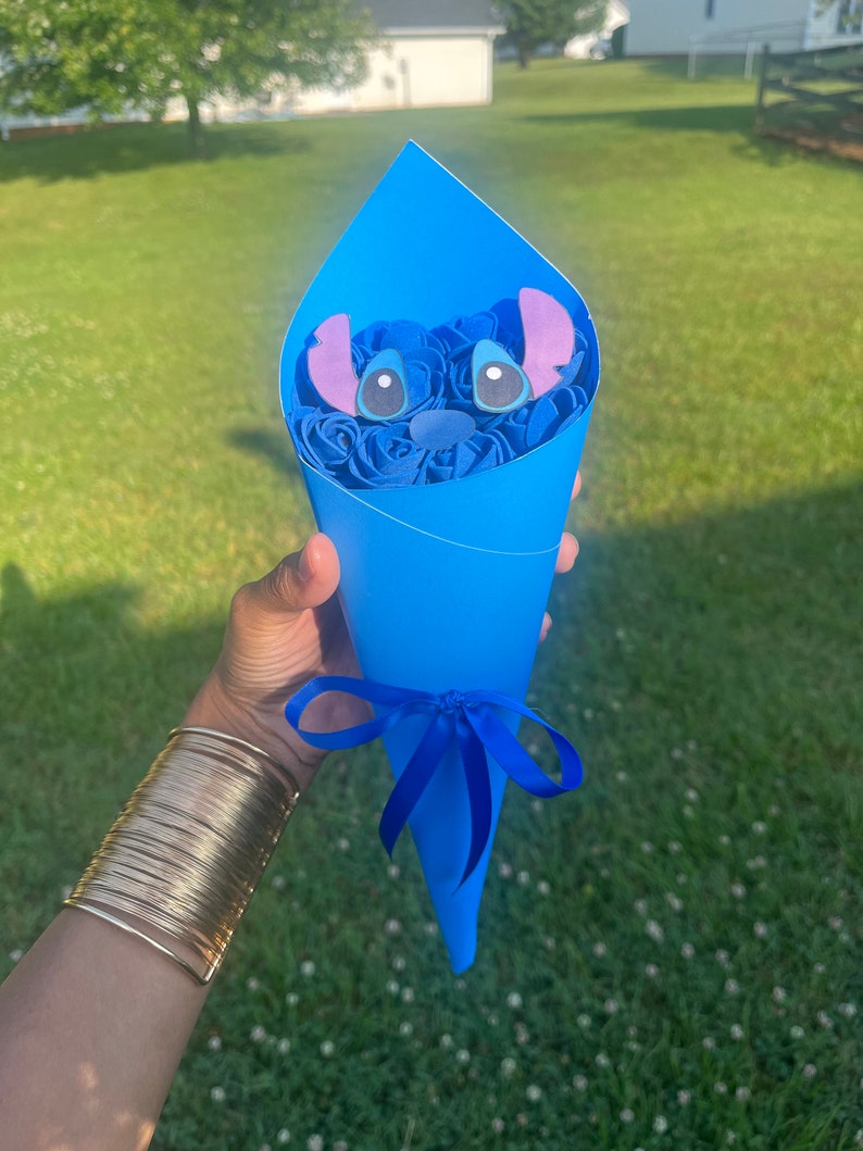 May include: A blue paper cone bouquet featuring a Stitch character face and blue roses. The bouquet is tied with a blue ribbon. The Stitch face has large eyes and purple ears. The background is a green lawn.