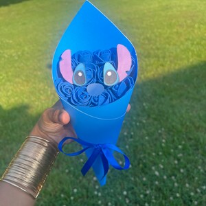 May include: A blue paper cone bouquet featuring blue paper roses and a Stitch character face. The bouquet is tied with a blue ribbon. The background is a green lawn.