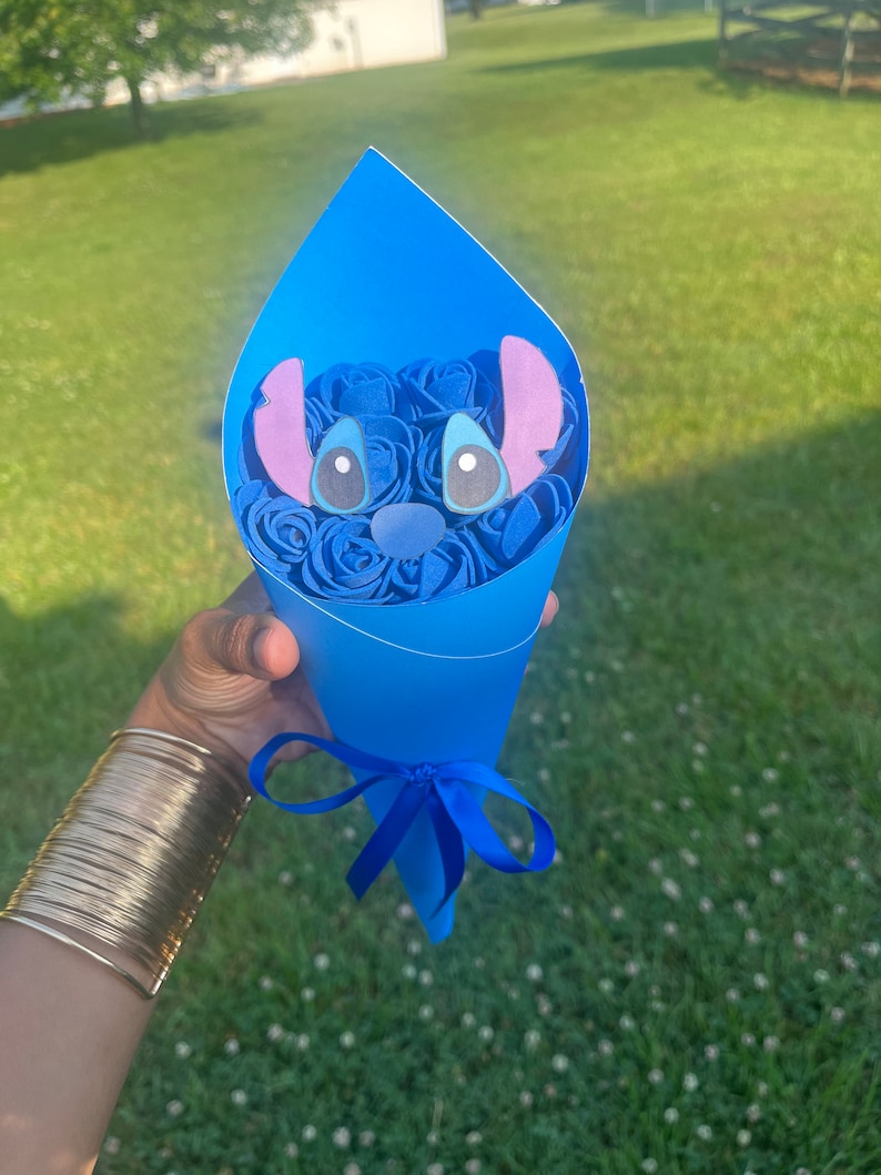May include: A blue paper cone bouquet featuring blue paper roses and a Stitch character face. The bouquet is tied with a blue ribbon. The background is a green lawn.