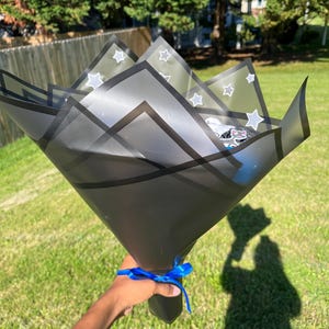 May include: A black gift bouquet wrapped in translucent paper with white star patterns. The bouquet is tied with a blue ribbon and held by a person's hand against a backdrop of green grass and trees.