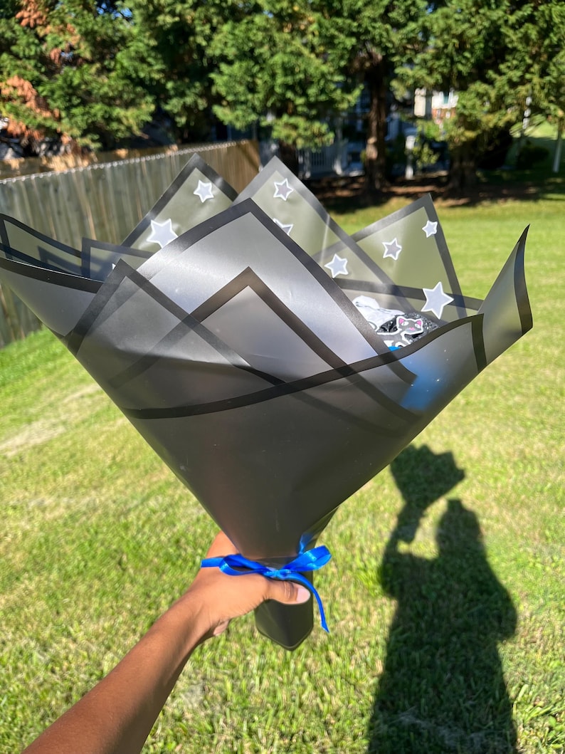 May include: A black gift bouquet wrapped in translucent paper with white star patterns. The bouquet is tied with a blue ribbon and held by a person's hand against a backdrop of green grass and trees.