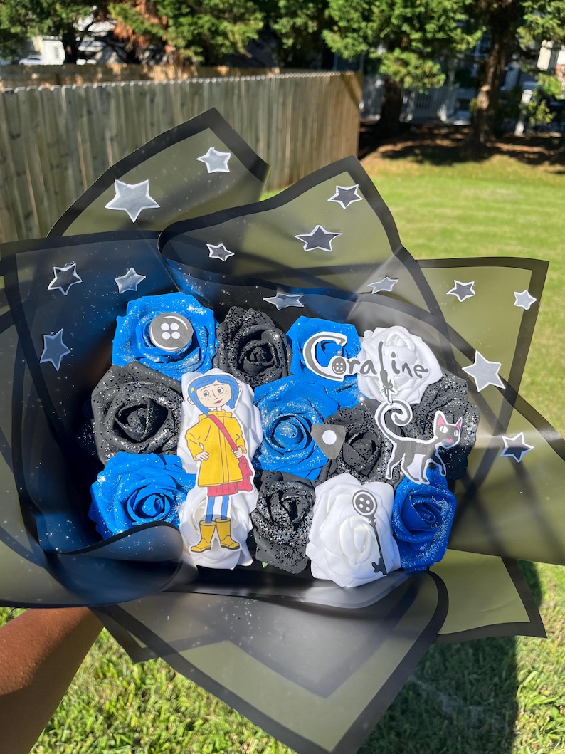 May include: A bouquet of blue, black, and white artificial roses, with a decorative wrapping featuring star patterns. The bouquet includes character cutouts and a button accent, creating a themed floral arrangement.