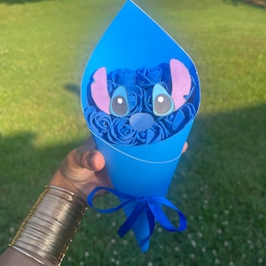 May include: A blue paper cone bouquet featuring blue roses and a cartoon character face. The bouquet is tied with a blue ribbon. The character has large eyes, a blue nose, and large ears.
