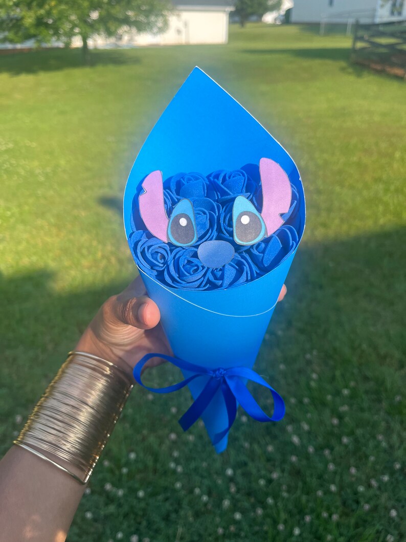 May include: A blue paper cone bouquet featuring blue roses and a cartoon character face. The bouquet is tied with a blue ribbon. The character has large eyes, a blue nose, and large ears.