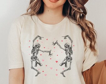 May include: A white t-shirt with a graphic of two skeletons dancing with pink hearts. The skeletons are wearing a casual pose with their arms outstretched. The shirt is being worn by a person with long brown hair and a brown hat.