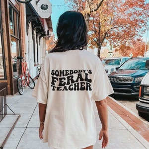 Peut inclure: Un t-shirt blanc surdimensionn&eacute; avec le texte "Somebody's Feral Teacher" imprim&eacute; en noir au dos. Le t-shirt est port&eacute; par une personne qui marche sur un trottoir.