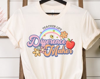 Op de afbeelding: Een crèmekleurig T-shirt met een regenboog, bloemen en een bij-ontwerp. De tekst op het shirt luidt "Teacher Life Difference Maker".