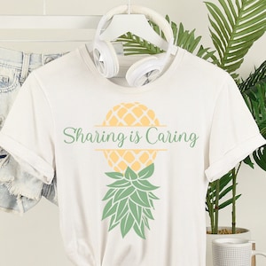 Puede incluir: Camiseta blanca con un gr&aacute;fico de pi&ntilde;a amarilla y el texto "Sharing is Caring" impreso en la parte delantera.