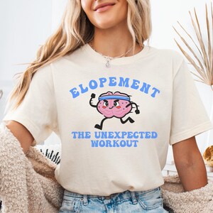 May include: A cream-coloured t-shirt with the text "ELOPEMENT THE UNEXPECTED WORKOUT" in blue. The shirt features a cartoon brain character with a headband, running. The model is wearing denim shorts and a beige cardigan.