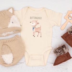 May include: A beige baby outfit set featuring a fox hat, diaper cover, and a bodysuit with a deer illustration. Includes brown fur-lined boots and a wooden rattle. The bodysuit has the text "Rutton Puck".
