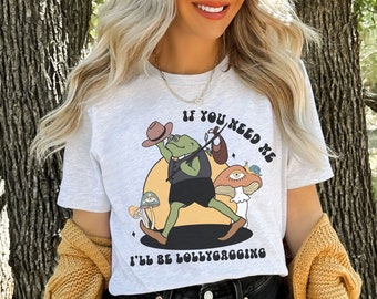 Op de afbeelding: Een wit T-shirt met een cartoon kikker die een hoed draagt en een muziekinstrument vasthoudt. De kikker is omringd door paddenstoelen en de tekst "Als je me nodig hebt, ben ik aan het lummelen".