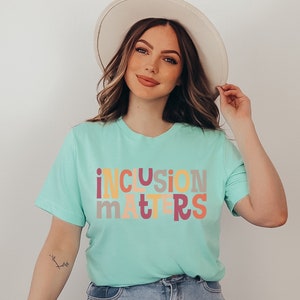 Puede incluir: Una mujer lleva una camiseta azul claro con el texto "inclusion matters" en letras de colores. Las letras son de diferentes colores, incluyendo rosa, amarillo, naranja y gris. Lleva un sombrero blanco y unos vaqueros azules.