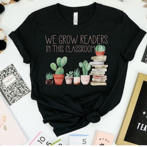 Może przedstawiać: Czarny T-shirt z napisem "We Grow Readers In This Classroom" w różowych literach. Projekt zawiera stos książek i rząd doniczkowych kaktusów.