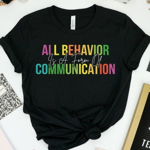 Op de afbeelding: Zwart T-shirt met de tekst "ALL BEHAVIOR Is A Form Of COMMUNICATION" in regenboogkleuren.