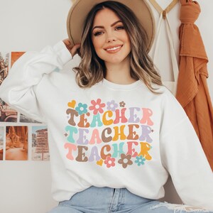 May include: White sweatshirt with a colorful retro-style graphic that says "Teacher Teacher Teacher" with flowers and hearts.
