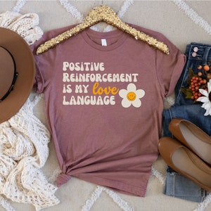 Puede incluir: Una camiseta color malva con el texto "POSITIVE REINFORCEMENT IS MY LOVE LANGUAGE" en una fuente retro. Una margarita blanca con una carita sonriente amarilla est&aacute; a la derecha del texto.