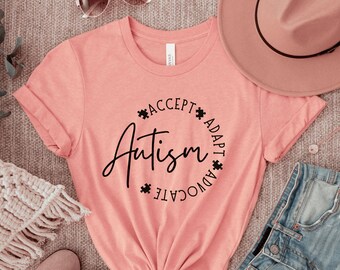 Puede incluir: Una camiseta rosa con un gráfico circular negro que dice "Autismo" con las palabras "Aceptar", "Adaptar" y "Abogar" que rodean la palabra "Autismo".