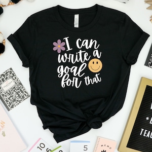 Puede incluir: Camiseta negra con texto blanco que dice "Puedo escribir un objetivo para eso" con una flor morada y una cara sonriente amarilla.