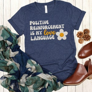 Puede incluir: Una camiseta azul marino con el texto "POSITIVE REINFORCEMENT IS MY love LANGUAGE" en una fuente retro con una margarita amarilla con una cara sonriente.