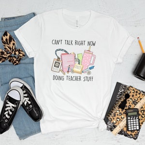 May include: White t-shirt with the text "Can't talk right now doing teacher stuff" and a graphic of school supplies, including a coffee mug, pencils, and a notebook.