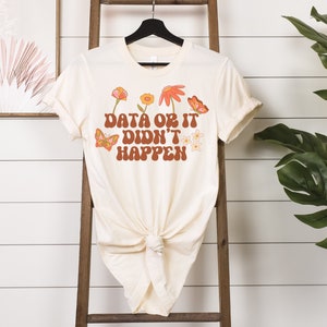 May include: A cream-colored t-shirt with a retro-style graphic that says "Data or it didn't happen". The graphic is surrounded by colorful flowers and butterflies.