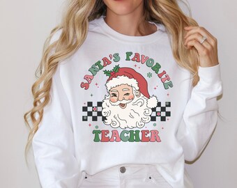 Könnte beinhalten: Weißer Pullover mit einem Retro-Grafik von Weihnachtsmann mit dem Text "Santa's Favorite Teacher" in Grün, Rot und Pink.