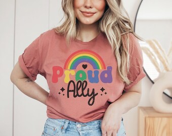 May include: A dusty rose t-shirt with rolled-up sleeves. The shirt features a rainbow arch design above the words "Proud Ally" in colorful letters. A small black heart and starbursts accent the text, promoting inclusivity and support.