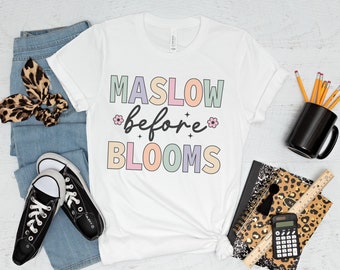 May include: White t-shirt with the text "MASLOW before BLOOMS" in colorful letters with a flower graphic. The shirt is laid out on a white surface with jeans, sneakers, pencils, a notebook, a calculator, and a mug.