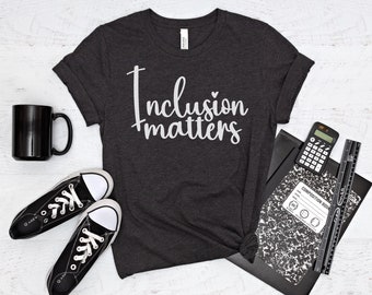 以下が含まれることがあります： 白い文字で「Inclusion matters」と書かれた濃いグレーのTシャツ。白い背景に黒いマグカップ、白黒の靴2足、黒いコンポジションノート、電卓、定規が置かれている。