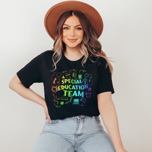 May include: A woman wearing a black t-shirt with a rainbow design that says "Special Education Team" surrounded by school-themed icons. She is wearing a brown hat and blue jeans.