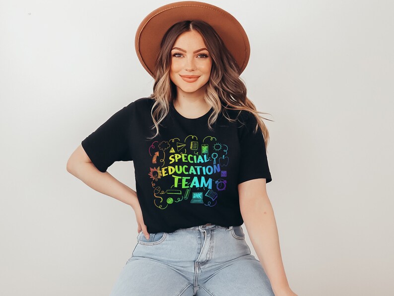 May include: A woman wearing a black t-shirt with a rainbow design that says "Special Education Team" surrounded by school-themed icons. She is wearing a brown hat and blue jeans.