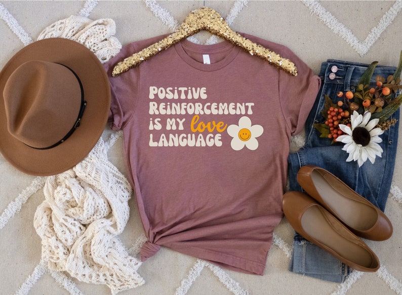 Puede incluir: Una camiseta color malva con el texto "POSITIVE REINFORCEMENT IS MY LOVE LANGUAGE" en una fuente retro. Una margarita blanca con una carita sonriente amarilla est&aacute; a la derecha del texto.