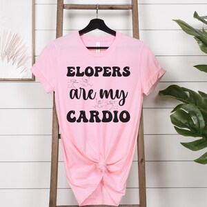 May include: Pink t-shirt with black text that reads "Elopers are my cardio". The text is in a retro font style. There are stick figure drawings of people running around the text.