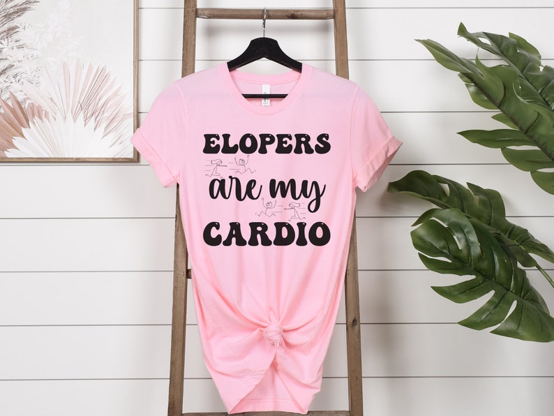 May include: Pink t-shirt with black text that reads "Elopers are my cardio". The text is in a retro font style. There are stick figure drawings of people running around the text.