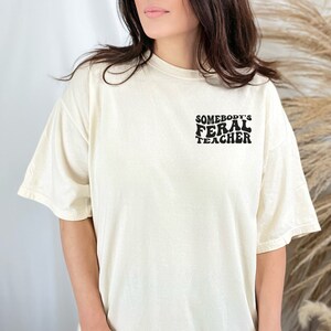 Peut inclure: Un t-shirt blanc avec un motif noir qui dit "Somebody's Feral Teacher" dans une police r&eacute;tro.