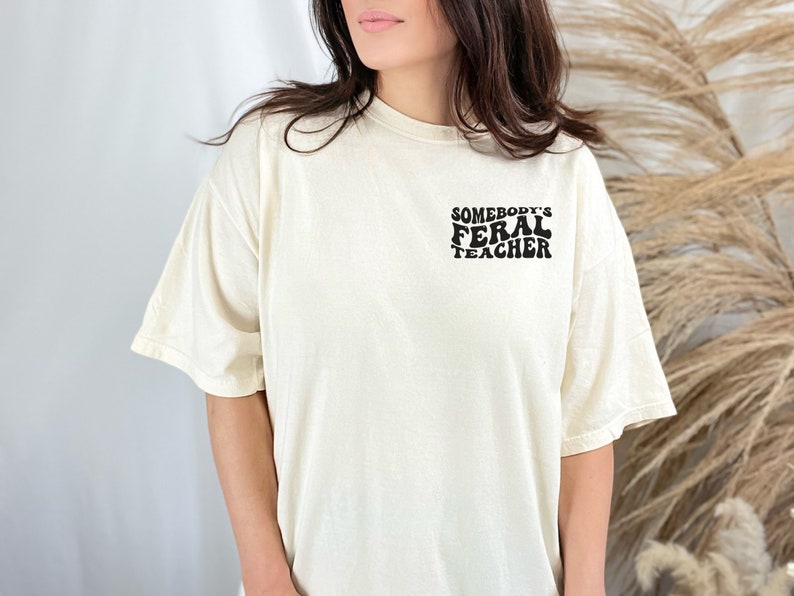 Peut inclure: Un t-shirt blanc avec un motif noir qui dit "Somebody's Feral Teacher" dans une police r&eacute;tro.