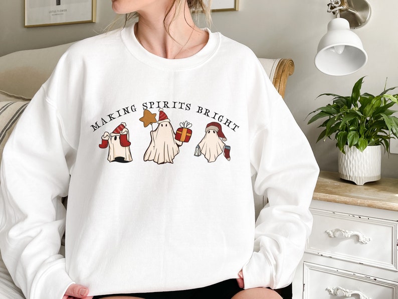 May include: White sweatshirt with a Christmas-themed design featuring three ghosts wearing Santa hats and holding holiday items. The text "Making Spirits Bright" is printed above the ghosts.
