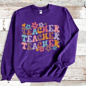 May include: Purple sweatshirt with the text "TEACHER" repeated three times in a retro font with colorful flowers and hearts.