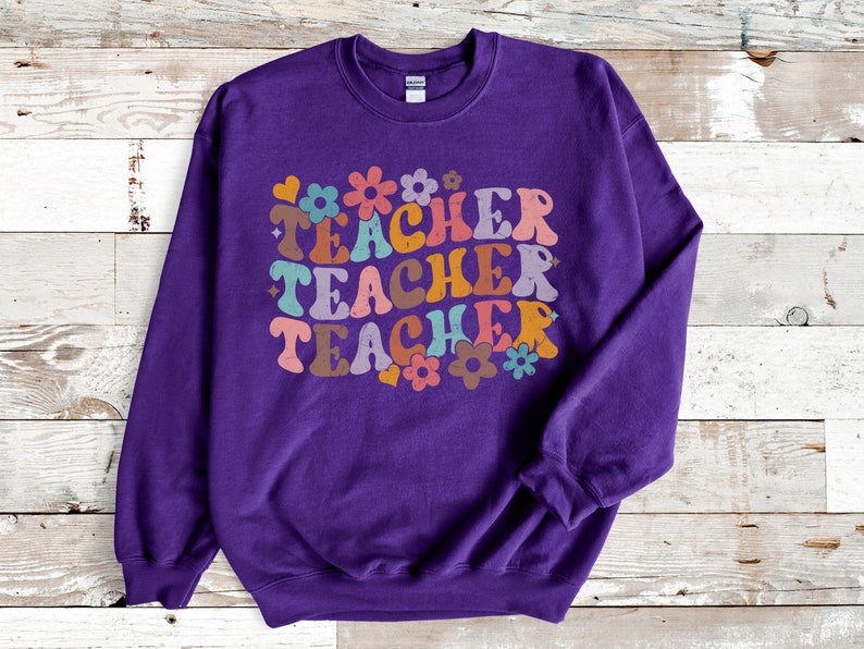 May include: Purple sweatshirt with the text "TEACHER" repeated three times in a retro font with colorful flowers and hearts.