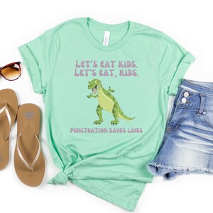 May include: A light green t-shirt featuring a cartoon dinosaur and the text "Let's eat kids. Let's eat, kids. Punctuation saves lives."