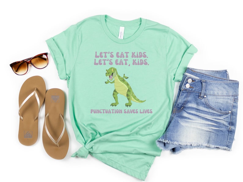 May include: A light green t-shirt featuring a cartoon dinosaur and the text "Let's eat kids. Let's eat, kids. Punctuation saves lives."